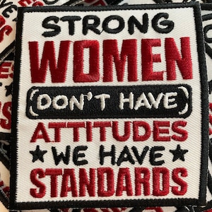 May include: Square white patch with black and red embroidered text. The text reads "STRONG WOMEN DON'T HAVE ATTITUDES WE HAVE STANDARDS". The patch has a black border.