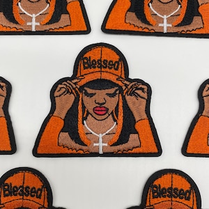 May include: An embroidered patch featuring a person with black and orange hair wearing an orange cap with the word Blessed embroidered in black. The person wears an orange long-sleeve top and a silver-toned necklace with a cross pendant.