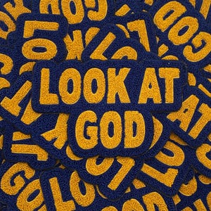 Op de afbeelding: Een verzameling marineblauwe patches met gouden letters. De centrale patch leest "LOOK AT GOD" in een vetgedrukte, getextureerde lettertype. Andere patches met het woord "GOD" zijn zichtbaar op de achtergrond, waardoor een gelaagd effect ontstaat.
