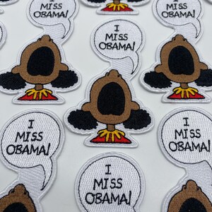 May include: A collection of embroidered patches featuring a cartoon character with a speech bubble that says "I Miss Obama". The character has brown skin, black hair, and a red shirt.