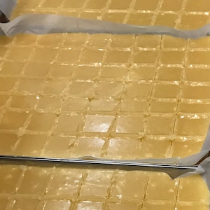 Op de afbeelding: Meerdere bakken met goudgele fudge, gesneden in vierkanten. De fudge zit in metalen bakken die zijn bekleed met bakpapier.
