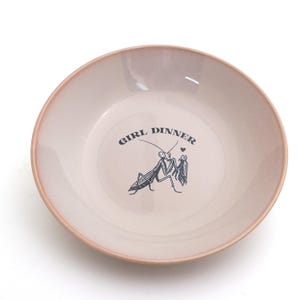 Peut inclure: Un bol rond en c&eacute;ramique rose clair avec un bord rose plus fonc&eacute;. Le bol porte l'inscription "GIRL DINNER" et une illustration noire de deux mantes religieuses. Un petit c&oelig;ur est au-dessus des mantes.