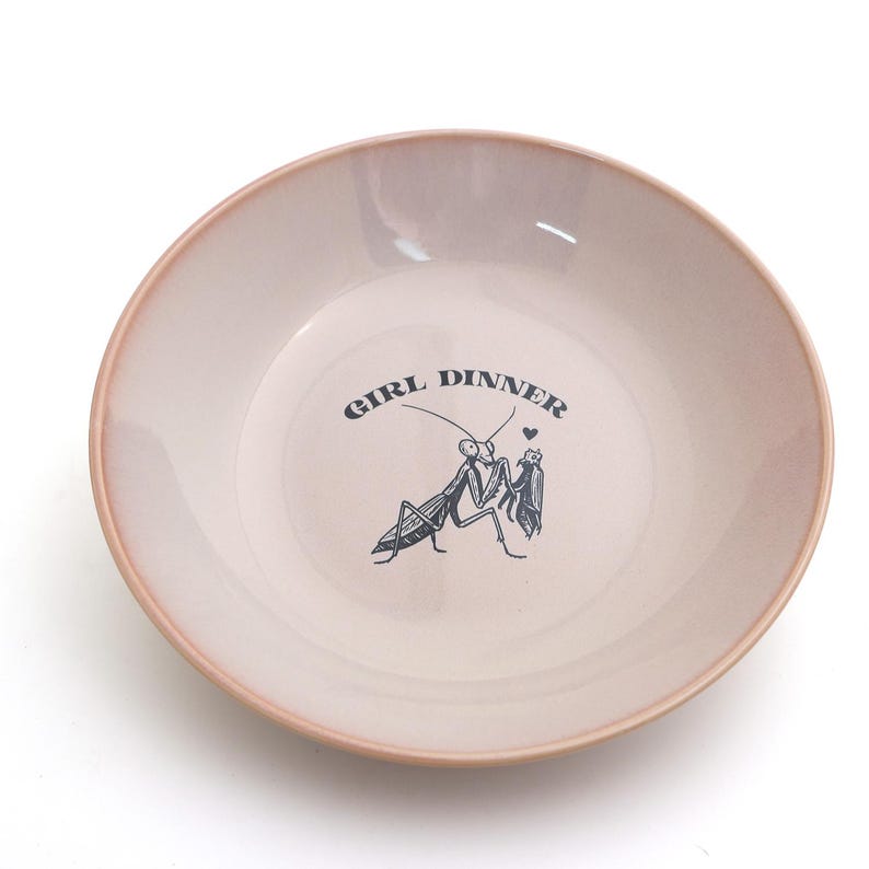 Peut inclure: Un bol rond en c&eacute;ramique rose clair avec un bord rose plus fonc&eacute;. Le bol porte l'inscription "GIRL DINNER" et une illustration noire de deux mantes religieuses. Un petit c&oelig;ur est au-dessus des mantes.