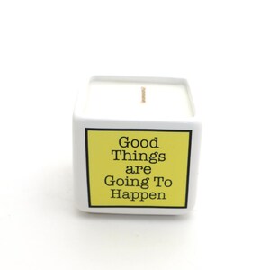 May include: A white square candle with a yellow label that reads "Good Things are Going To Happen" in black text. The candle has a single wick and is set against a white background.
