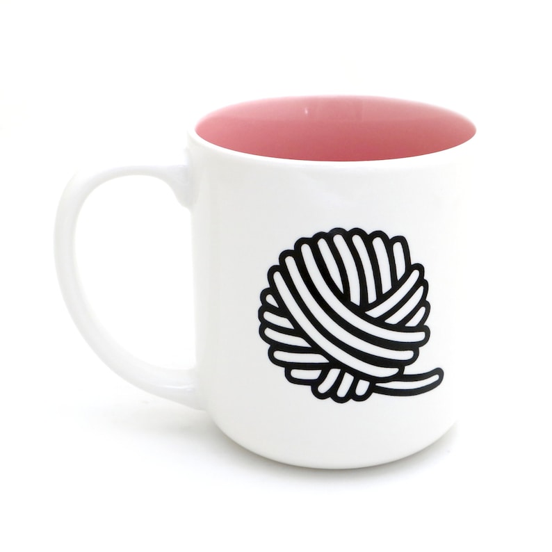 Op de afbeelding: Witte keramische mok met een roze binnenkant en een zwart-witte illustratie van een bol garen aan de voorkant.