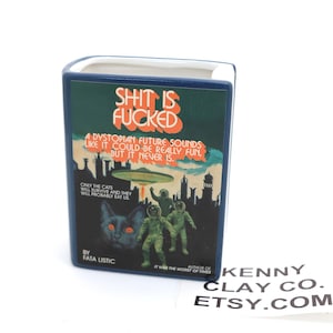 May include: A blue ceramic bookend shaped like a book with the title "Shit is Fucked: A Dystopian Future Sounds Like It Could Be Really Fun, But It Never Is." The cover features a black cat with red eyes, three astronauts, and a flying saucer. The text "By Fata Listic" is at the bottom of the cover.