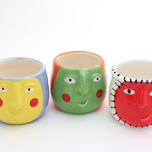 May include: Three ceramic face planters in a row. The first is yellow with a blue rim, the second is green with orange hair, and the third is red with white stitching. Each has a smiling face.