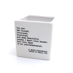 May include: White ceramic square planter with a black typewriter font quote from F. Scott Fitzgerald's letter to Zelda in 1959. The quote reads: "You are the finest, loveliest, tenderest, and most beautiful person I have ever known- and even that is an understatement."