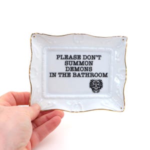 May include: White ceramic trinket dish with a gold rim and ornate detailing. The dish features the text "PLEASE DON'T SUMMON DEMONS IN THE BATHROOM" in black, along with a skull graphic. A decorative and humorous bathroom accessory.