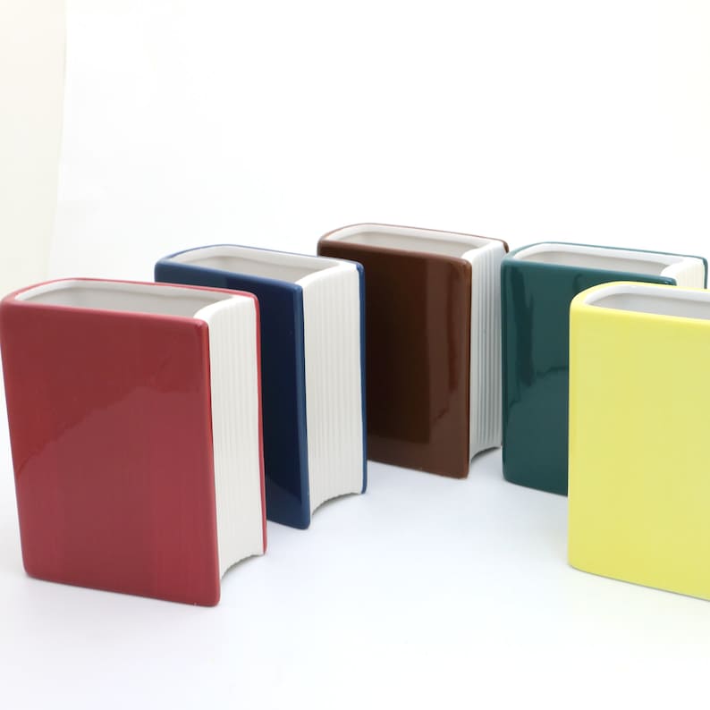 May include: Five ceramic book-shaped planters in different colors: red, navy blue, brown, green, and yellow. The planters have white pages on the spine.