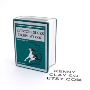 May include: A teal blue ceramic bookend shaped like a book with the title "Everyone Sucks Except My Dog" and a cartoon dog illustration. The bookend is a handmade item from Kenny Clay Co. Etsy.com.