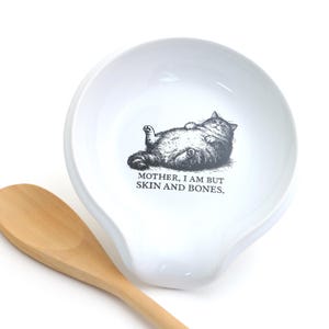 May include: A white ceramic spoon rest featuring a black and white illustration of a plump cat lying on its back. Below the cat, the text reads, MOTHER, I AM BUT SKIN AND BONES. A light-colored wooden spoon is positioned next to the spoon rest.