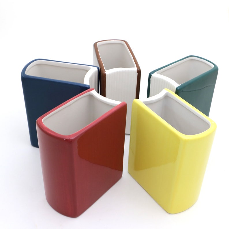 May include: Five ceramic book-shaped planters in different colors, including red, yellow, blue, green, and brown. The planters are arranged in a circle, with the openings facing up.