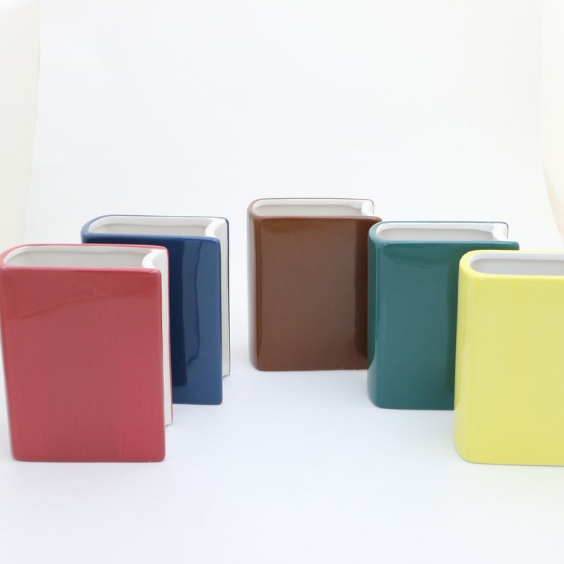 May include: Five ceramic book-shaped planters in different colors: red, blue, brown, teal, and yellow. The planters are arranged in a row on a white surface.