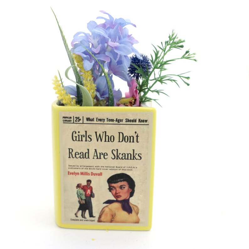 May include: A yellow ceramic planter shaped like a book with the title "Girls Who Don't Read Are Skanks" and a vintage illustration of a woman on the cover. The planter is filled with a small arrangement of blue, yellow, and green flowers.