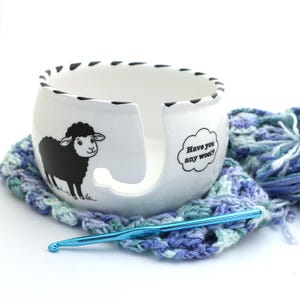 May include: A white ceramic yarn bowl with a black sheep illustration and the text "Have you any wool?" A blue crochet hook rests on a blue, green, and white crocheted square. The bowl has a curved opening for yarn.