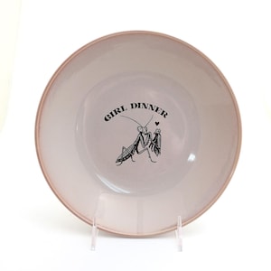 Peut inclure: Une assiette ronde en c&eacute;ramique blanc cass&eacute; avec un bord rose. L'assiette porte l'inscription "GIRL DINNER" dans une police incurv&eacute;e, ainsi qu'une illustration noire de deux insectes. L'assiette est pr&eacute;sent&eacute;e sur un support transparent.