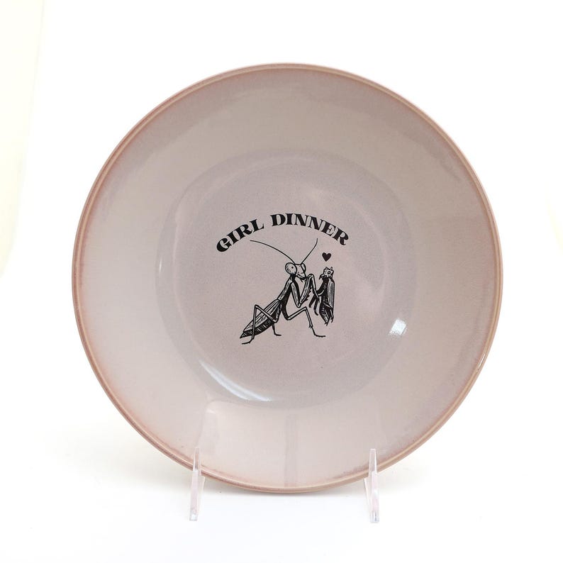 Peut inclure: Une assiette ronde en c&eacute;ramique blanc cass&eacute; avec un bord rose. L'assiette porte l'inscription "GIRL DINNER" dans une police incurv&eacute;e, ainsi qu'une illustration noire de deux insectes. L'assiette est pr&eacute;sent&eacute;e sur un support transparent.
