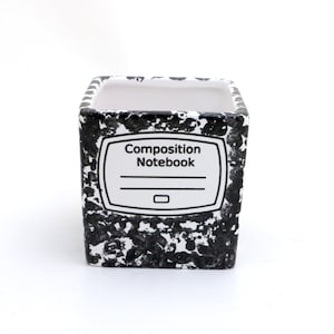 May include: Black and white ceramic planter with a composition notebook design. The planter has a square shape and a white rim. The text "Composition Notebook" is printed on the front of the planter.