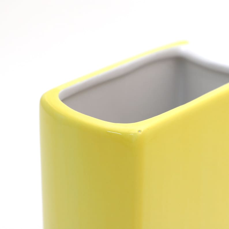 May include: A yellow ceramic vase with a rectangular shape and rounded edges. The vase is empty and has a white interior.