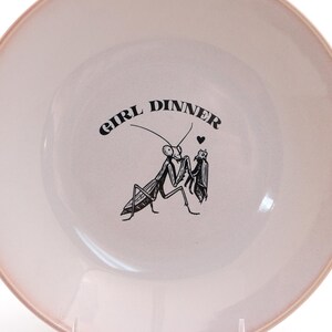 Peut inclure: Une assiette ronde en c&eacute;ramique rose clair avec une illustration noire de deux mantes religieuses et les mots "GIRL DINNER". L'assiette a une surface l&eacute;g&egrave;rement textur&eacute;e et un bord rose plus fonc&eacute;.