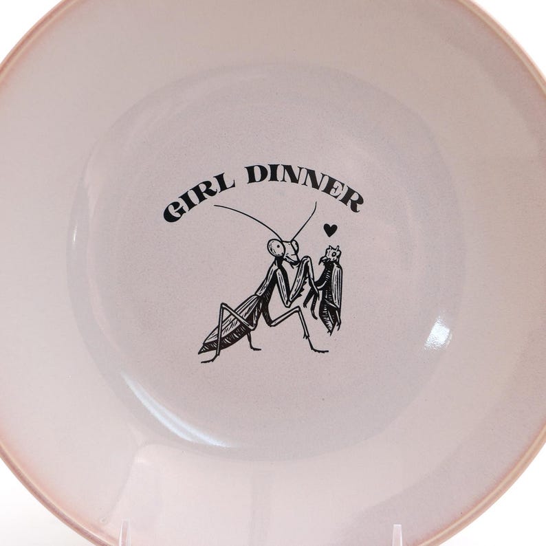 Peut inclure: Une assiette ronde en c&eacute;ramique rose clair avec une illustration noire de deux mantes religieuses et les mots "GIRL DINNER". L'assiette a une surface l&eacute;g&egrave;rement textur&eacute;e et un bord rose plus fonc&eacute;.