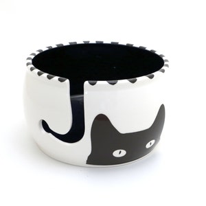 May include: White ceramic yarn bowl with a black cat design. The cat's face is on the front of the bowl, and its tail wraps around the back. The bowl has a hole in the center for yarn to be dispensed.