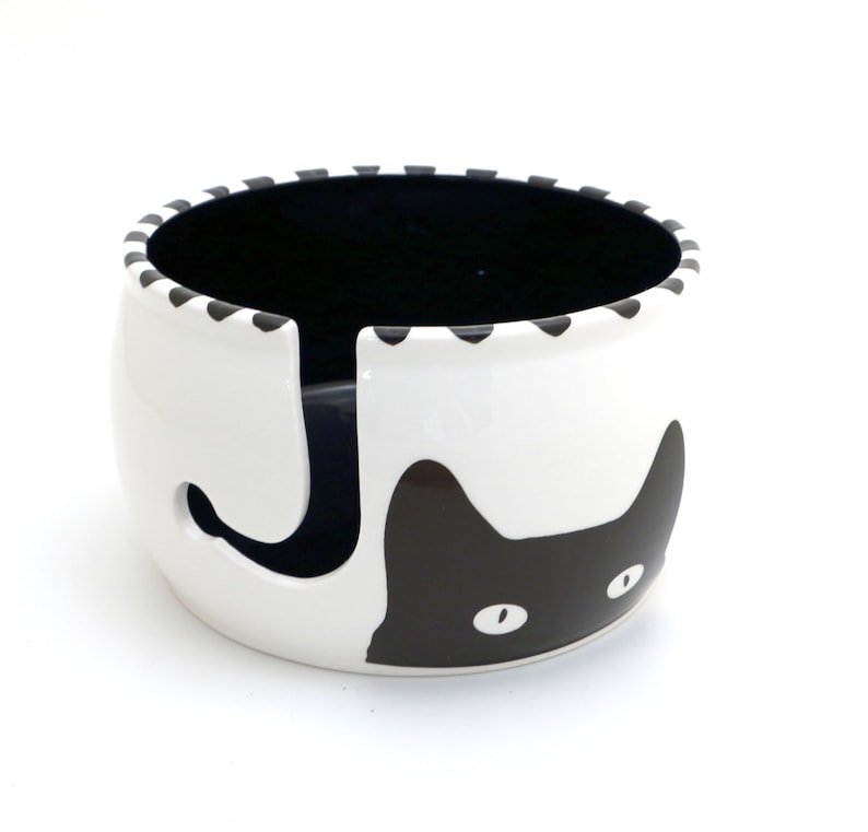 May include: White ceramic yarn bowl with a black cat design. The cat's face is on the front of the bowl, and its tail wraps around the back. The bowl has a hole in the center for yarn to be dispensed.