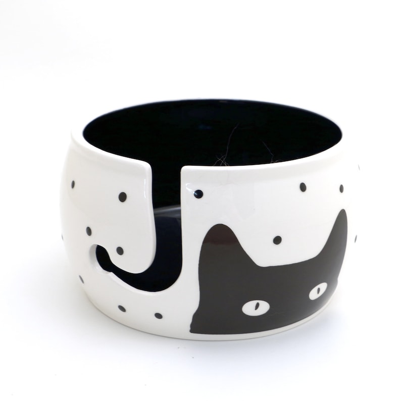 May include: White ceramic yarn bowl with a black cat design and black polka dots. The bowl has a hole in the side for yarn to feed through.