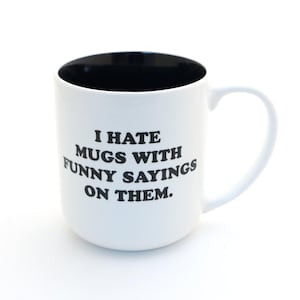 May include: White ceramic mug with a black interior and a curved handle. The mug features the text "I HATE MUGS WITH FUNNY SAYINGS ON THEM" in black, bold letters. The mug is designed for hot beverages.