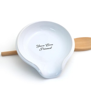May include: White ceramic spoon rest with the text "You've Been Poisoned" in black script. The spoon rest has a curved shape with a raised edge and a small handle rest. A wooden spoon rests on the side.