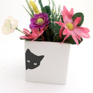 May include: White ceramic planter with a black cat silhouette peeking out from the side. The planter is filled with a variety of artificial flowers, including pink, white, and purple.
