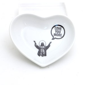 May include: White heart-shaped ceramic dish with a black and white image of a person with arms outstretched and a speech bubble that says "Love You More".