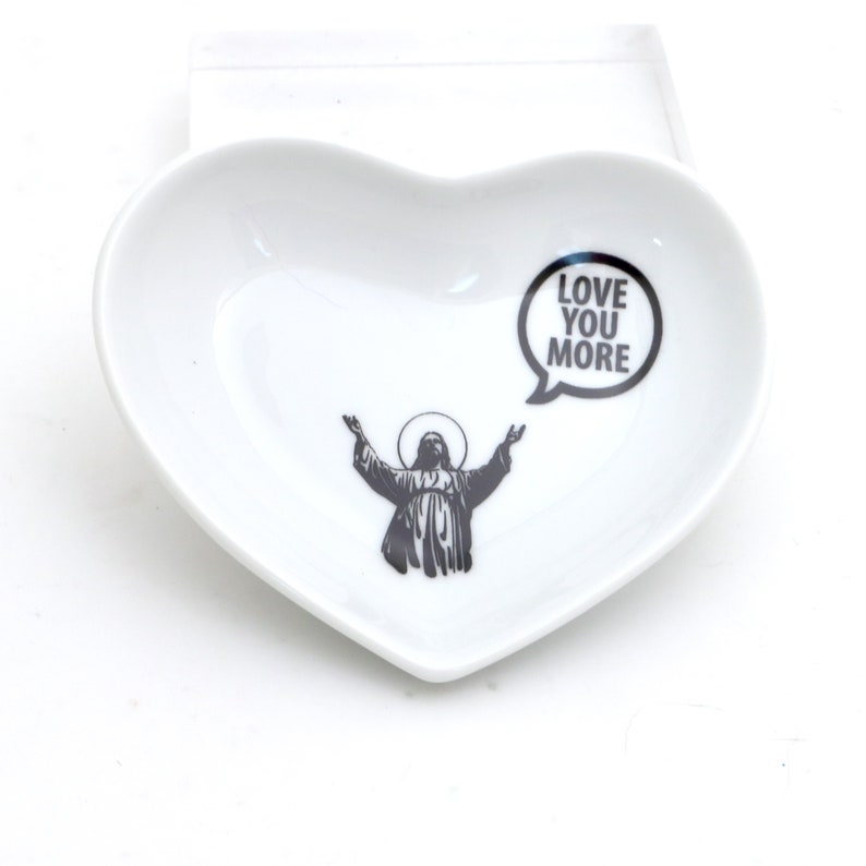 May include: White heart-shaped ceramic dish with a black and white image of a person with arms outstretched and a speech bubble that says "Love You More".
