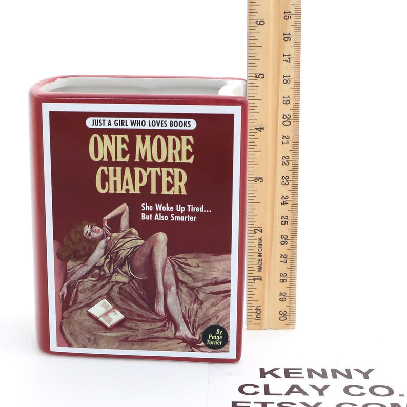 May include: A red book-shaped ceramic pencil holder with the title "One More Chapter" and the text "Just a girl who loves books. She woke up tired... But also smarter. By Paige Turner." The holder is 15.2 cm tall.