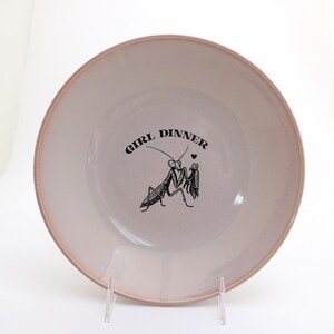 Peut inclure: Une assiette ronde en c&eacute;ramique rose clair avec un bord rose plus fonc&eacute;. L'assiette pr&eacute;sente une illustration noire de deux insectes et les mots "GIRL DINNER" en texte noir.