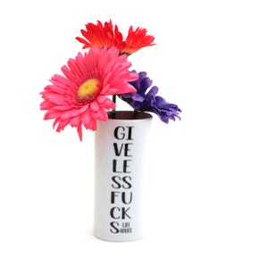 May include: A white cylindrical vase featuring the black text "GIVE LESS FUCKS LIFE ADVICE" arranged vertically. The vase contains a bouquet of artificial flowers, including large pink gerbera daisies, red flowers, and a purple flower.