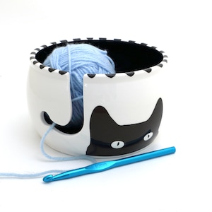 May include: A white ceramic yarn bowl with a black cat design. The bowl has a hole in the side for yarn to feed through. A blue ball of yarn is inside the bowl. A blue crochet hook is lying on a white surface.