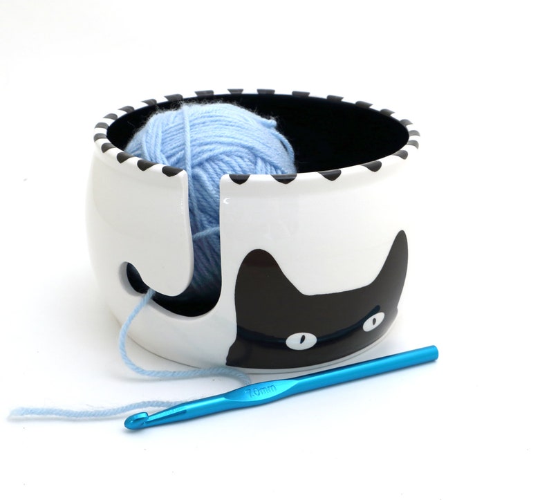 May include: A white ceramic yarn bowl with a black cat design. The bowl has a hole in the side for yarn to feed through. A blue ball of yarn is inside the bowl. A blue crochet hook is lying on a white surface.