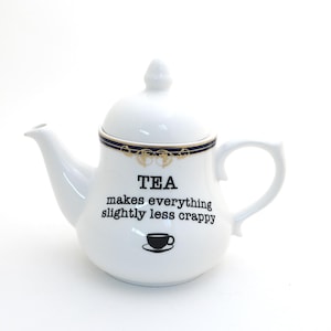 May include: White ceramic teapot with a lid and handle. The teapot has a black and gold rim design and the text "TEA makes everything slightly less crappy" with a teacup graphic.