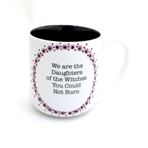 Może przedstawiać: Biały kubek ceramiczny z czarnym rantem i uchwytem. Kubek ma wz&oacute;r w fioletowe i czerwone kropki wok&oacute;ł krawędzi i napis "We are the Daughters of the Witches You Could Not Burn" w kolorze czarnym.
