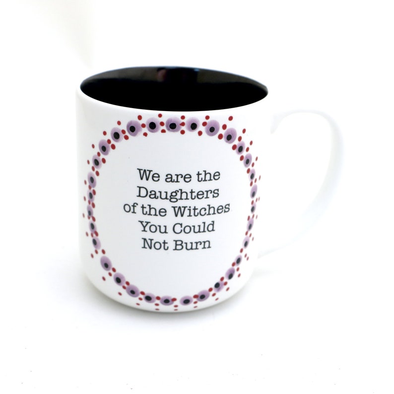 Może przedstawiać: Biały kubek ceramiczny z czarnym rantem i uchwytem. Kubek ma wz&oacute;r w fioletowe i czerwone kropki wok&oacute;ł krawędzi i napis "We are the Daughters of the Witches You Could Not Burn" w kolorze czarnym.