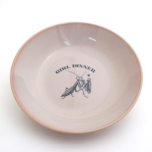 Peut inclure: Un bol en c&eacute;ramique rose clair avec un bord rose plus fonc&eacute;. Le bol pr&eacute;sente une illustration noire de deux mantes religieuses et les mots "GIRL DINNER" au-dessus. Un petit c&oelig;ur est au-dessus des mantes.