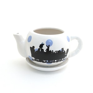 May include: A white ceramic teapot planter with a matching saucer, featuring light blue polka dots and a black silhouette graphic of characters seated at a table. The teapot has a curved handle, a short spout, and an open top for planting.