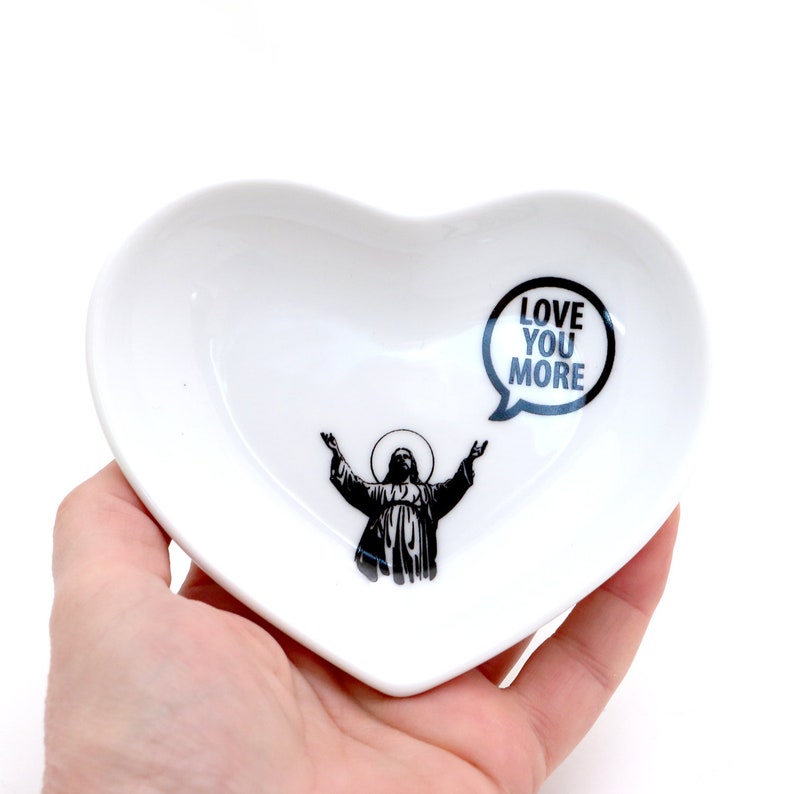 May include: White heart-shaped ceramic dish with a black silhouette of a person with arms outstretched and a speech bubble that says "Love You More".