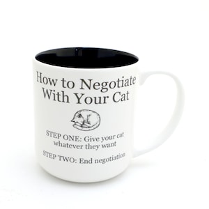 May include: White ceramic mug with a black interior. The mug has a humorous saying about negotiating with cats: "How to Negotiate With Your Cat. Step One: Give your cat whatever they want. Step Two: End negotiation."