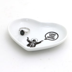 May include: White heart-shaped ceramic dish with a black and white image of a person with outstretched arms and the text "LOVE YOU MORE" in a speech bubble. A silver ring with a black stone is on the dish.
