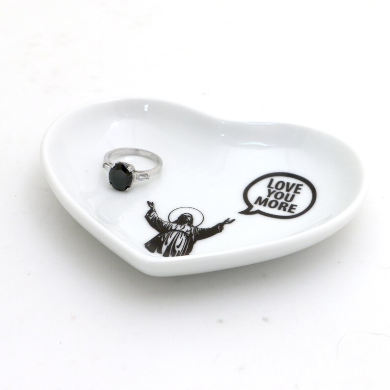 May include: White heart-shaped ceramic dish with a black and white image of a person with outstretched arms and the text "LOVE YOU MORE" in a speech bubble. A silver ring with a black stone is on the dish.