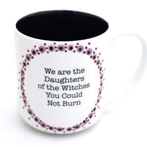 Może przedstawiać: Biały kubek ceramiczny z czarnym wnętrzem i wzorem z fioletowych i czerwonych kropek wok&oacute;ł krawędzi. Kubek ma napis "We are the Daughters of the Witches You Could Not Burn" nadrukowany czarnym kolorem.