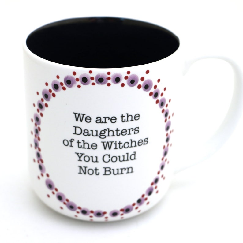 Może przedstawiać: Biały kubek ceramiczny z czarnym wnętrzem i wzorem z fioletowych i czerwonych kropek wok&oacute;ł krawędzi. Kubek ma napis "We are the Daughters of the Witches You Could Not Burn" nadrukowany czarnym kolorem.